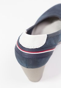 Modrá semišová lodička na vysokém podpatku s bílým a červeným pruhovaným lemováním a bílou značkovou nášivkou na patě, na bílém pozadí.