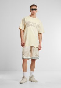Light yellow short-sleeve t-shirt with a large, patterned graphic. Beige shorts with a subtle design and green trim. White sneakers and sunglasses.