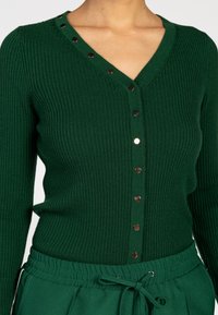 Close-up van een persoon die een donkergroene geribbelde knopen cardigan en bijpassende groene broek met trekkoord draagt.