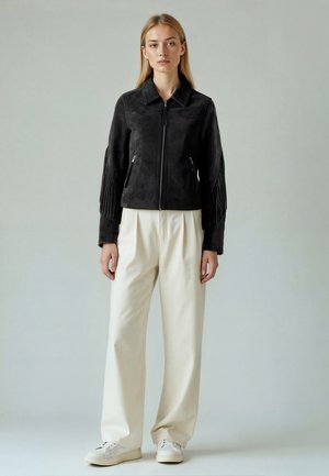 Nő fekete velúr bojtokkal díszített dzsekit, fehér bőszárú nadrágot és fehér tornacipőt visel, egyszerű világosszürke háttér előtt áll.