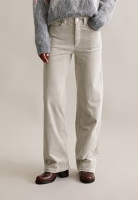 Helle beige Cordhosen mit geradem Schnitt, hoher Taille, Fronttaschen und kontrastierenden Nähten, kombiniert mit braunen Stiefeln mit klobigem Absatz.