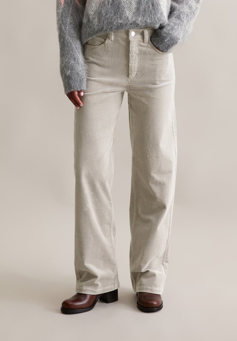 Helle beige Cordhosen mit geradem Schnitt, hoher Taille, Fronttaschen und kontrastierenden Nähten, kombiniert mit braunen Stiefeln mit klobigem Absatz.