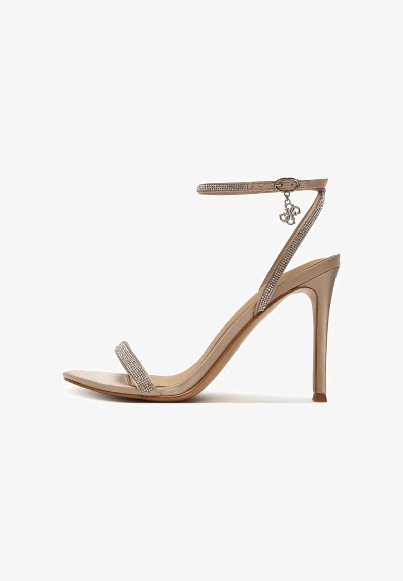 Sandale à talon haut beige avec bride à la cheville, bout ouvert, fines lanières ornées de strass, et un petit charme métallique suspendu à la boucle.