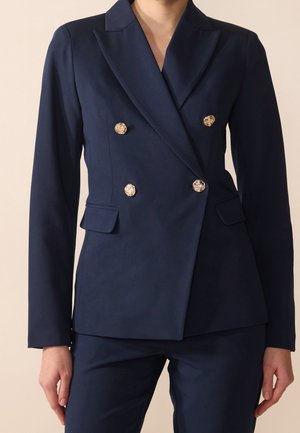 Femme portant un blazer croisé bleu marine sur mesure avec quatre boutons décoratifs dorés et un pantalon assorti bleu marine.