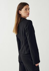 Melnā pielāgotā žakete ar garām piedurknēm, gludu faktūru, strukturētu piegriezumu un vienu aizmugurējo venti. Minimalistisks dizains bez dekorācijām.