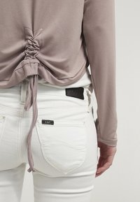 Jasnofioletowa bluzka z długim rękawem i marszczonym tyłem, zestawiona z białymi dżinsami z czarną skórzaną metką i kieszeniami. Gładka tekstura.