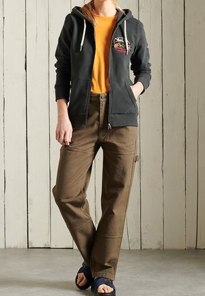 Grijze hoodie met rits en trekkoorden, oranje T-shirt, bruine loszittende broek en donkere sandalen tegen een houten achtergrond.