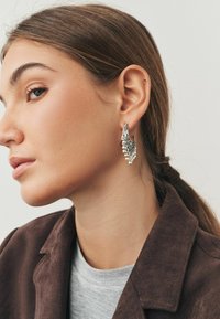 Sølvøreringe med detaljeret udsmykning, der har et buet design og hængende perler. Modellen bærer en brun jakke over en grå skjorte.