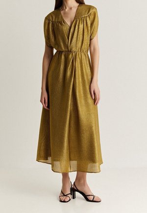 Gold, pleated maxi dress with a deep V-neck and short puff sleeves. The fabric has a textured sheen. Paired with black strappy heels.