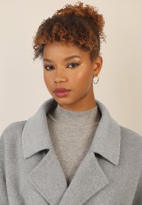 Cappotto grigio in lana con colletto a punte indossato sopra un maglione grigio a collo alto aderente. Presenta una texture morbida e una silhouette elegante.