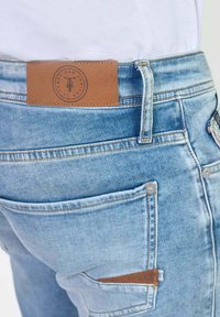 Vaqueros de denim azul claro con un parche de cuero marrón en la cinturilla, que presentan costuras en contraste y detalles en los bolsillos laterales.