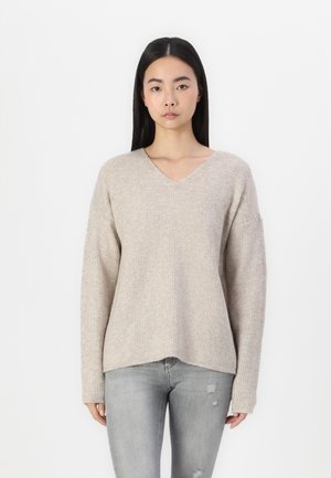 Beige knitted sweater with a V-neck, dropped shoulders, and long sleeves. Soft texture and ribbed pattern. Paired with distressed light grey jeans.
