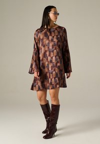 Langärmliges Kleid in Brauntönen mit geometrischem Muster. Kombiniert mit knielangen burgunderfarbenen Stiefeln, minimalem Schmuck und einem neutralen Hintergrund.