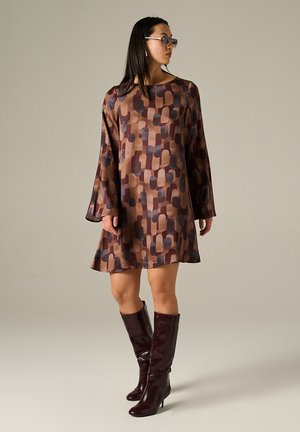 Abito a maniche lunghe in tonalità di marrone con un motivo geometrico. Abbinato a stivali borgogna al ginocchio, accessori minimali e uno sfondo neutro.