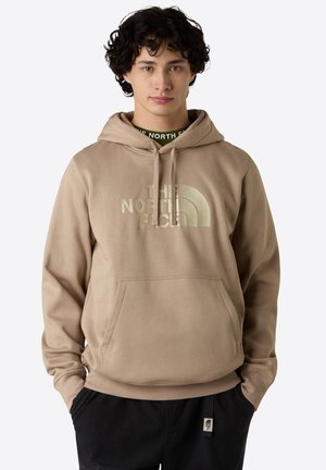 Sudadera beige hecha de material suave, con un bolsillo delantero y un logo blanco prominente. Los puños y el dobladillo acanalados ofrecen un ajuste ceñido.