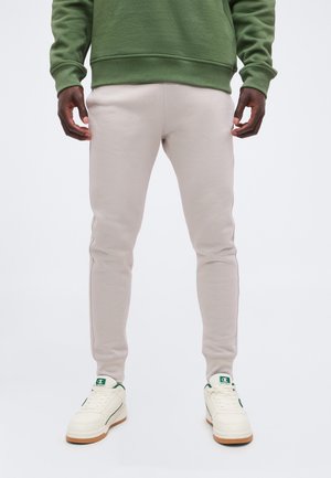 Hombre vistiendo pantalones jogger slim fit de color beige claro, zapatillas blancas con detalles verdes y una sudadera verde, de pie sobre un fondo blanco.