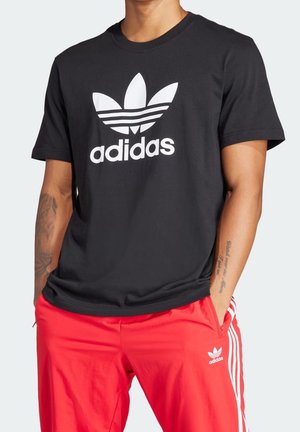 Homme portant un t-shirt Adidas noir avec logo blanc et un pantalon de survêtement Adidas rouge avec des bandes blanches, mains dans les poches, tatouages visibles sur les bras.