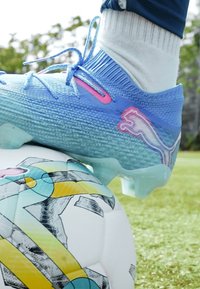 Botín de fútbol azul con parte superior texturizada, acentos en rosa y suela semitransparente, reposando sobre un balón de fútbol colorido y estampado en la hierba.