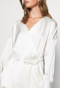 Blouse portefeuille en satin blanc avec un col en V profond, des manches longues et une ceinture nouée à la taille. Texture lisse avec une finition élégante.