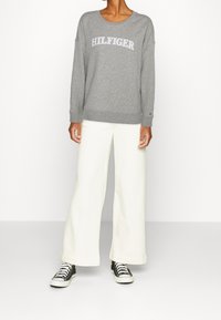 Persoon in een grijze "HILFIGER" sweatshirt, wijde witte broek en zwarte sneakers, staand tegen een effen witte achtergrond.