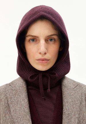Frau mit blauen Augen, die eine weinrote Strickkapuze mit Kordelzügen und einen beigen Tweedmantel trägt, schaut direkt in die Kamera.