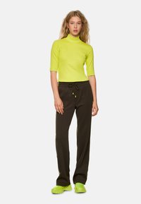 Neon yellow fitted top with a high neckline, paired with loose, dark brown pants featuring a drawstring waist and green accents. Chunky green shoes.