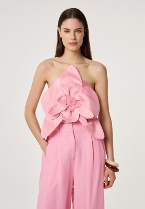 Vrouw draagt een strapless roze top met een groot bloemmotief en bijpassende roze hoge taille broek, staand met handen in de zakken.