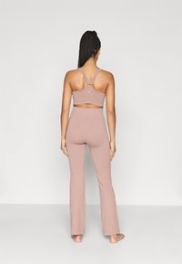 Σετ αθλητικών ρούχων σε απαλό ροζ χρώμα που περιλαμβάνει ένα κοντό τοπ με λουριά σε σχήμα σταυρού και κολάν με πλατειά παντελόνια. Λείο, ελαστικό υλικό με ελάχιστες λεπτομέρειες.