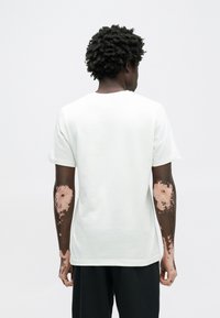 Balta trumpomis rankovėmis marškinėliai su apvaliomis kaklo iškirptėmis, minkšta medžiaga, laisvas siluetas, matomos odos atspalvių variacijos ant rankų.