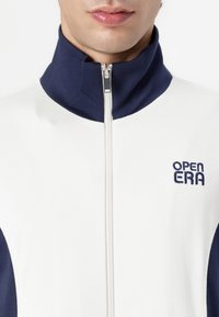 Veste zippée blanche avec col marine, présentant une fermeture éclair visible et un logo brodé "OPEN ERA" en bleu sur la zone de la poitrine.