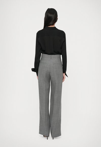 Femeie cu păr lung și închis la culoare, îmbrăcată într-o bluză neagră și pantaloni largi gri cu talie înaltă, stând cu fața spre un fundal alb.