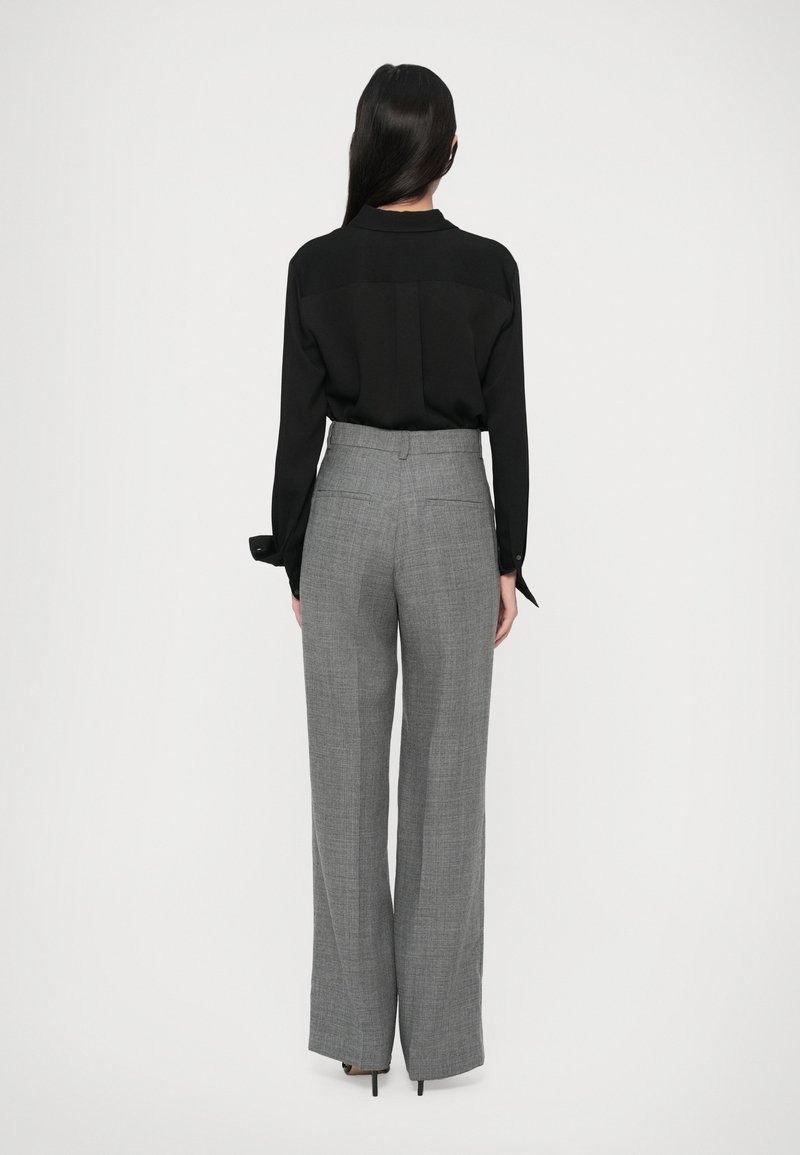 Donna con lunghi capelli scuri che indossa una blusa nera e pantaloni larghi grigi a vita alta, in piedi di fronte a uno sfondo bianco.