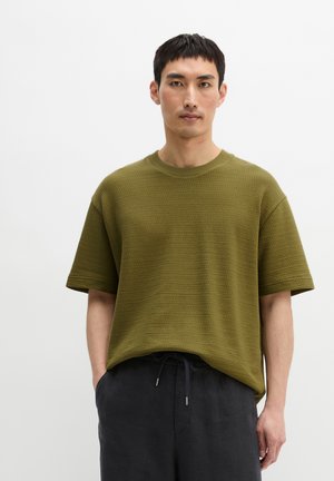 Mężczyzna ubrany w teksturowaną oliwkowozieloną koszulę z krótkim rękawem i czarne spodnie ze sznurkiem, stojący z rękami w kieszeniach na białym tle.