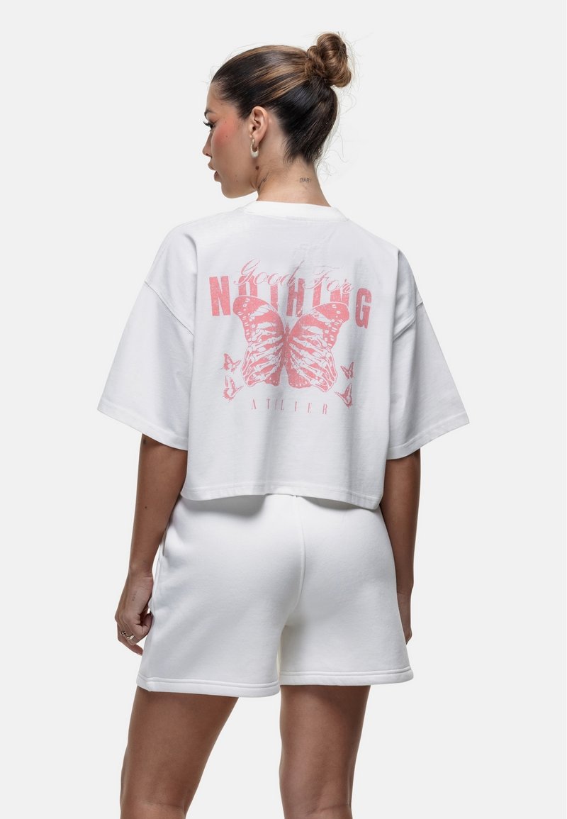 T-shirt bianca corta con maniche corte e grafica di una farfalla rosa sul retro, abbinata a pantaloni corti bianchi, per un look casual.