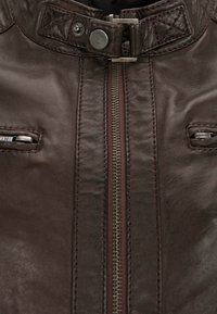 Bruine leren jas met hoge kraag, ritssluiting en twee ritszakken op de borst met accenten van zilverkleurig hardware. Geweven afwerking.