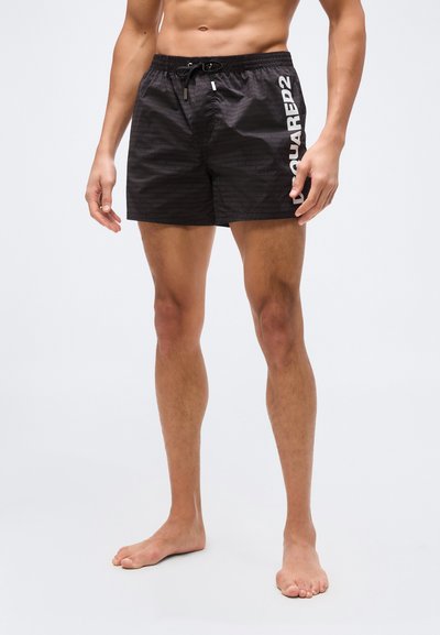 Personne musclée portant un short de bain noir avec cordon de serrage, debout pieds nus sur un fond uni. Texte "DSQUARED2" sur la jambe gauche.