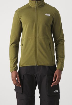 Mies, jolla on päällään oliivinvihreä vetoketjullinen takki ja mustat cargo-housut, molemmissa The North Face -logot rinnassa ja taskussa.