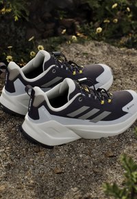 Paire de chaussures de trail Adidas Terrex noires et blanches avec des accents jaunes reposant sur du gravier avec de petites fleurs en arrière-plan.