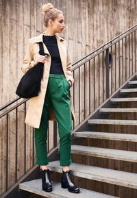 Beige trenchcoat, zwart topje, groene broek en zwarte enkellaarzen. Model staat op de trap met een zwarte tas, met een gladde textuur en een nauwsluitende pasvorm.