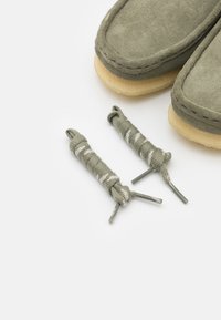 Mocassins de camurça verde com solas de cor creme, apresentando dois atacadores verdes-azeitona a condizer com riscas brancas.