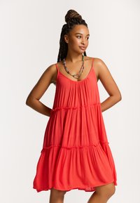 Rochie roșie fără mâneci, realizată dintr-un material ușor, cu straturi în trepte și accente subtile de volane. Modelul poartă un colier multicolor.