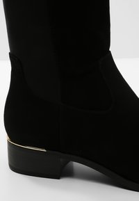 Black suede ankle boot featuring a sleek design, elastic side panels, and a gold-tone accent on the heel. Smooth texture and rounded toe.