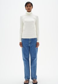 Weißer, gerippter Rollkragenpullover mit langen Ärmeln, kombiniert mit lässigen blauen Jeans. Das Outfit wird mit flachen schwarzen Sandalen abgerundet.