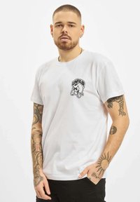 Camiseta de algodón blanca con un estampado gráfico negro que incluye la palabra "UNFAIR" en el lado izquierdo del pecho. Mangas cortas, ajuste casual.