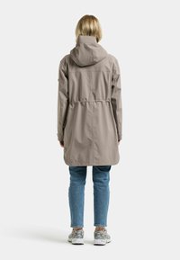 Vaalean beige sadetakki hupulla, kiristetyllä vyötäröllä ja pitkillä hihoilla. Yhdistetty sinisiin farkkuihin ja vaalean värisiin lenkkareihin.