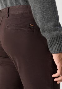 Gros plan d'une personne portant un pantalon marron foncé avec une petite étiquette orange "BOSS" sur la poche arrière et une manche de pull en maille grise.