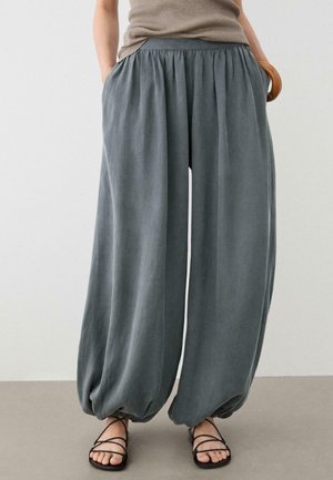 Frau trägt lockere, graue Ballonhosen mit elastischen Bündchen an den Knöcheln, ein beiges ärmelloses Oberteil und schwarze flache Riemchensandalen.