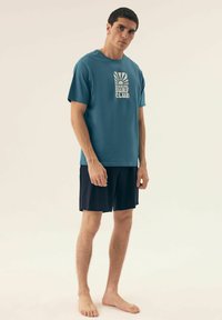 Turkusowa bawełniana koszulka z grafiką klubu surfowego, w połączeniu z ciemnymi szortami. Model stoi na boso, prezentując swobodny, casualowy wygląd.