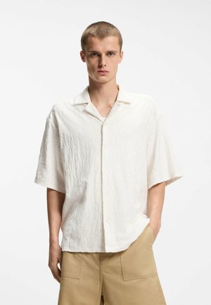 Ung mand med kort hår iført tekstureret hvid skjorte med korte ærmer og beige shorts, stående mod en ensartet hvid baggrund.