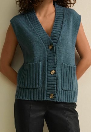 Gilet en tricot turquoise, avec un décolleté en V, trois boutons marron, des finitions côtelées et deux poches avant, associé à un pantalon en cuir noir.